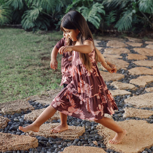 little girl wearing a long brown dress with tie sleeve with light pink lei awapuhi print