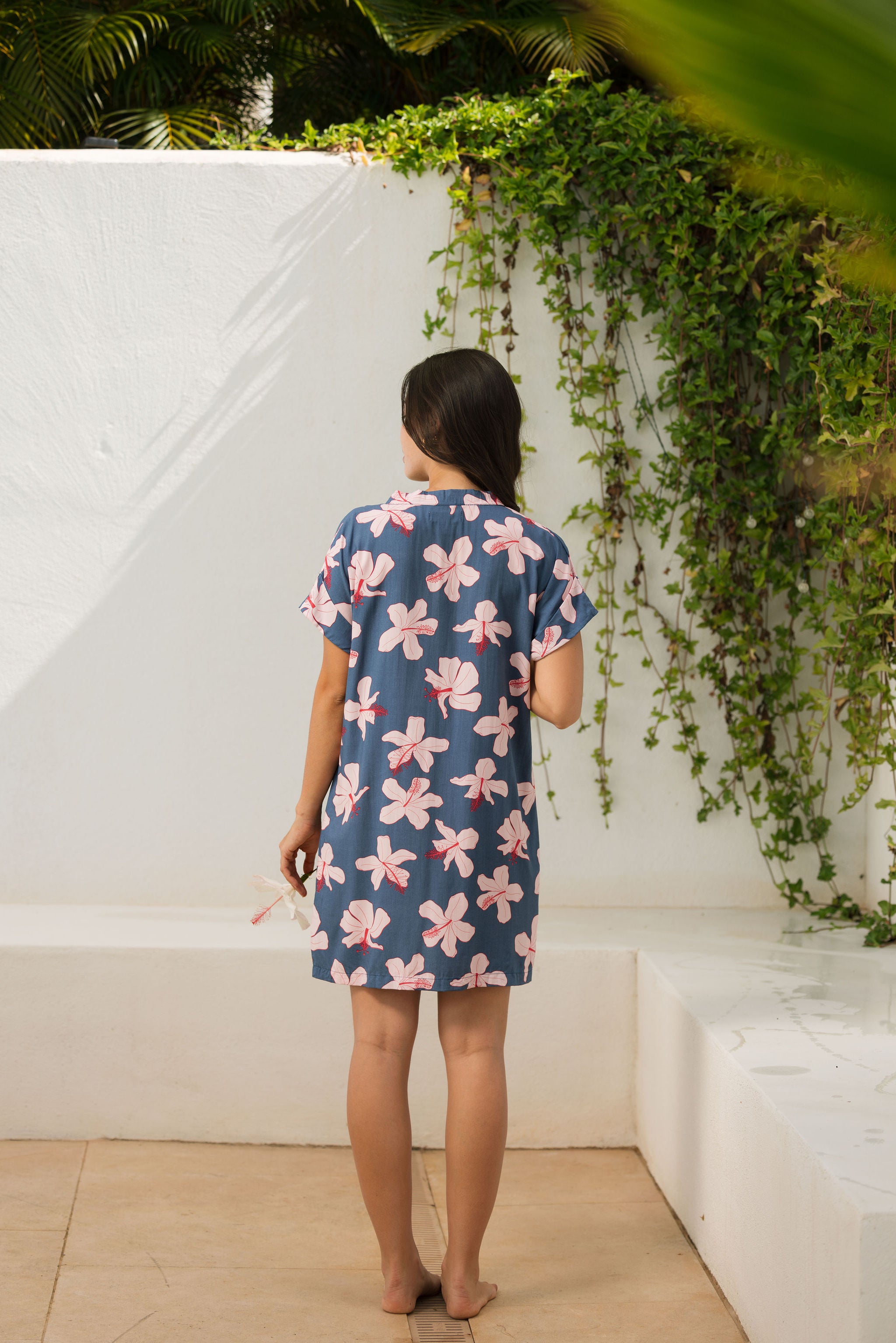 woman with black hair wearing a casual shirt dress with button up v neckline in a navy and pink hibiscus print.