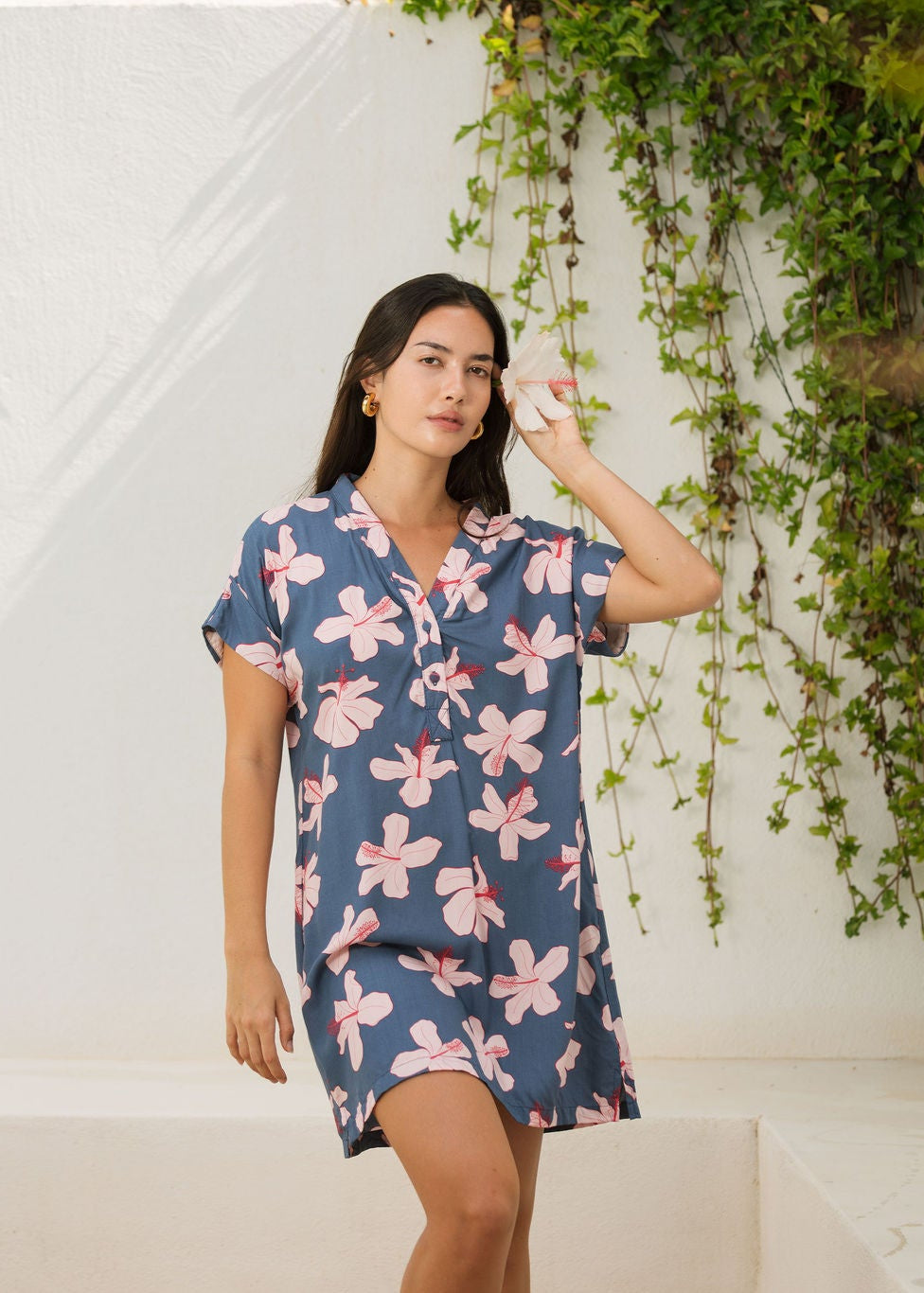 woman with black hair wearing a casual shirt dress with button up v neckline in a navy and pink hibiscus print.
