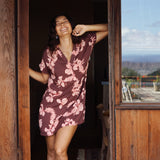 woman with brown hair wearing a brown colored button up v neckline dress with light pink lei awapuhi pattern
