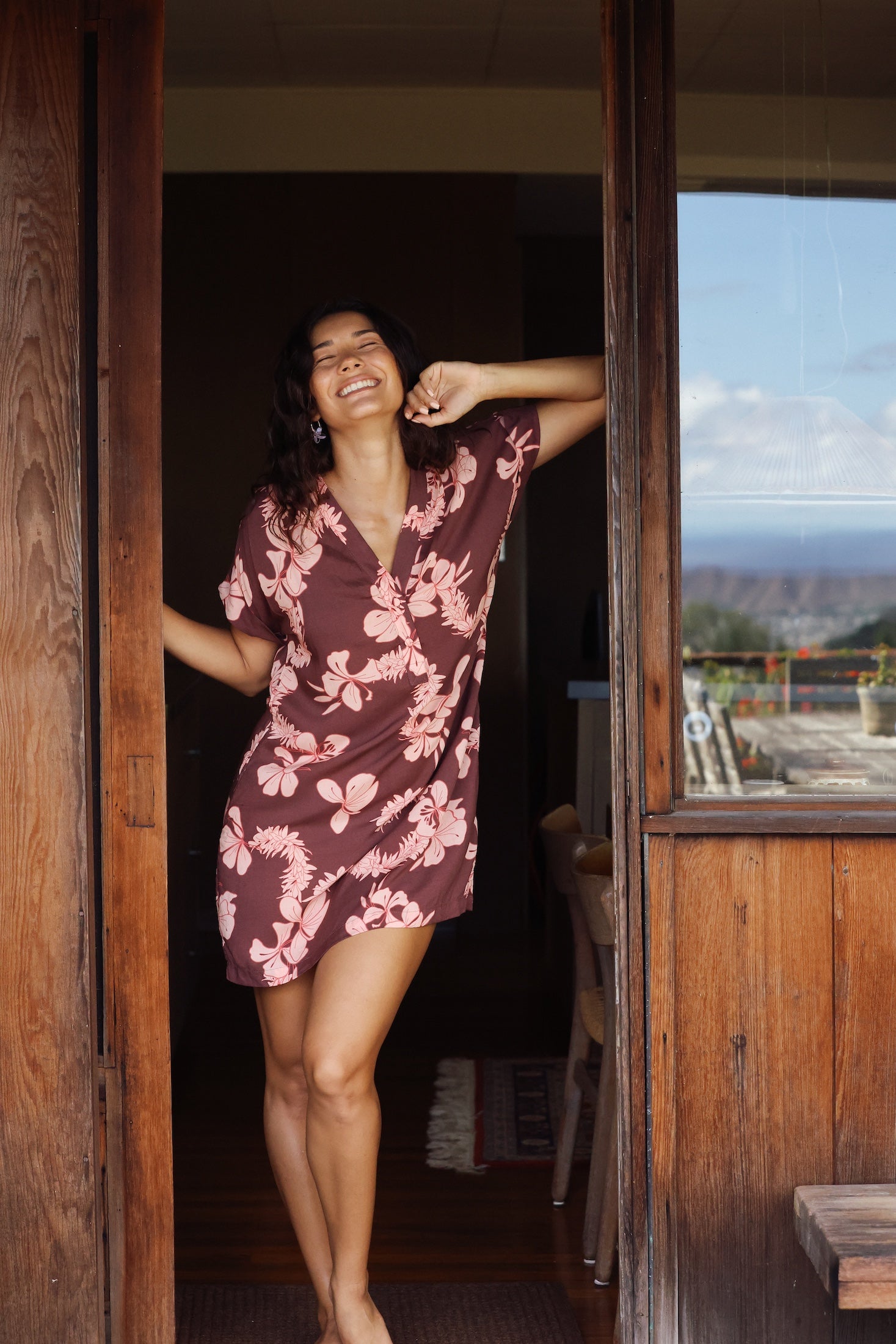 woman with brown hair wearing a brown colored button up v neckline dress with light pink lei awapuhi pattern