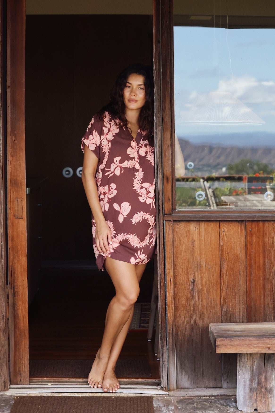 woman with brown hair wearing a brown colored button up v neckline dress with light pink lei awapuhi pattern