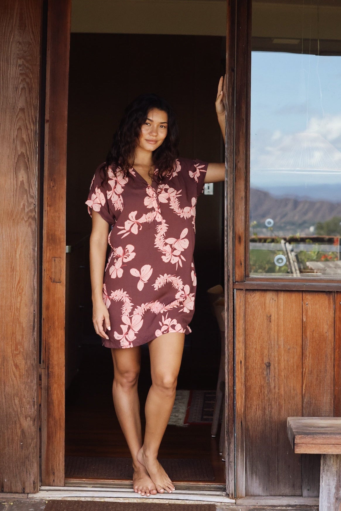 woman with brown hair wearing a brown colored button up v neckline dress with light pink lei awapuhi pattern