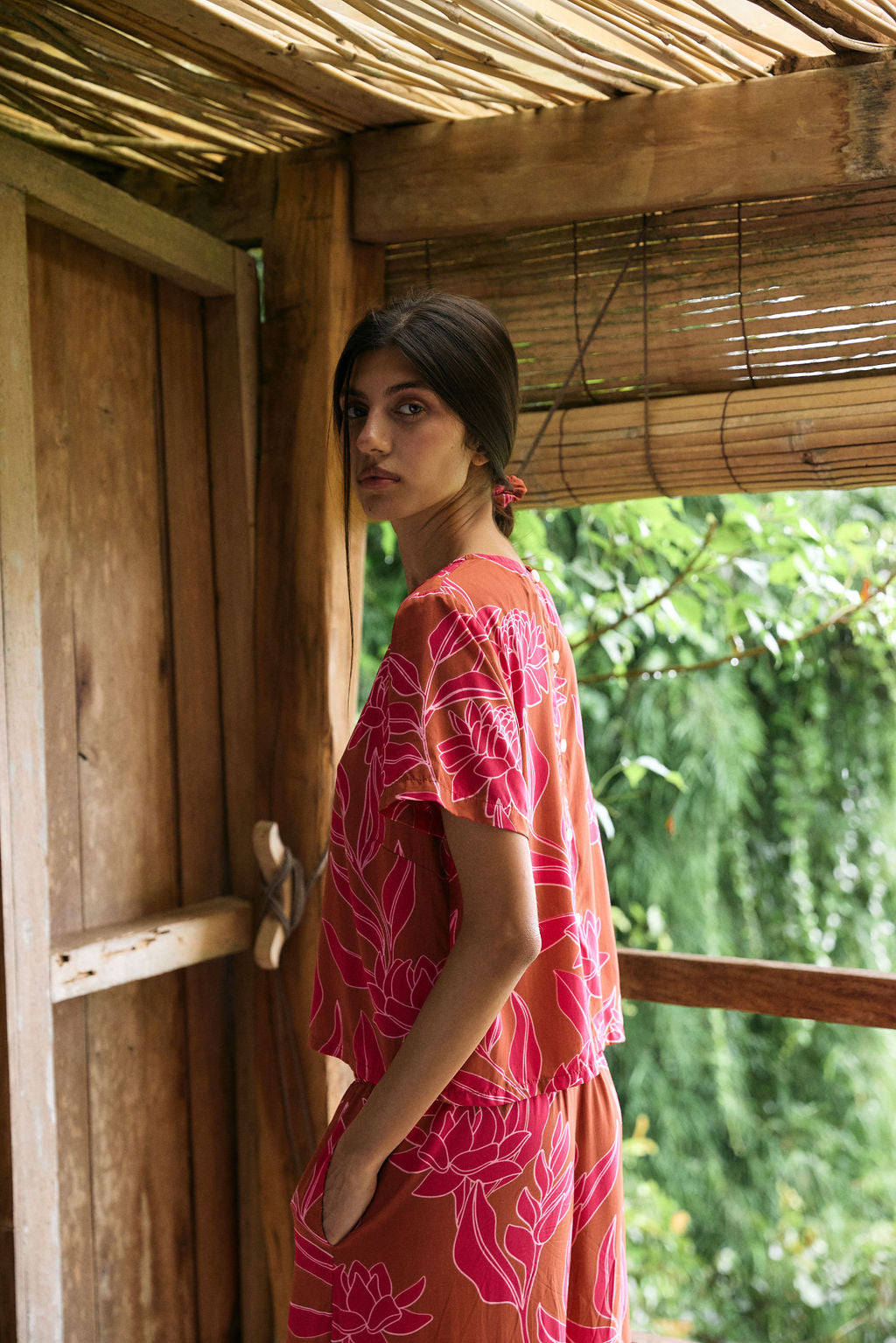 A woman with dark hair wears a YIREH t-shirt top with shell button back. It is the Noe Top in the Garden 'Awapuhi print.