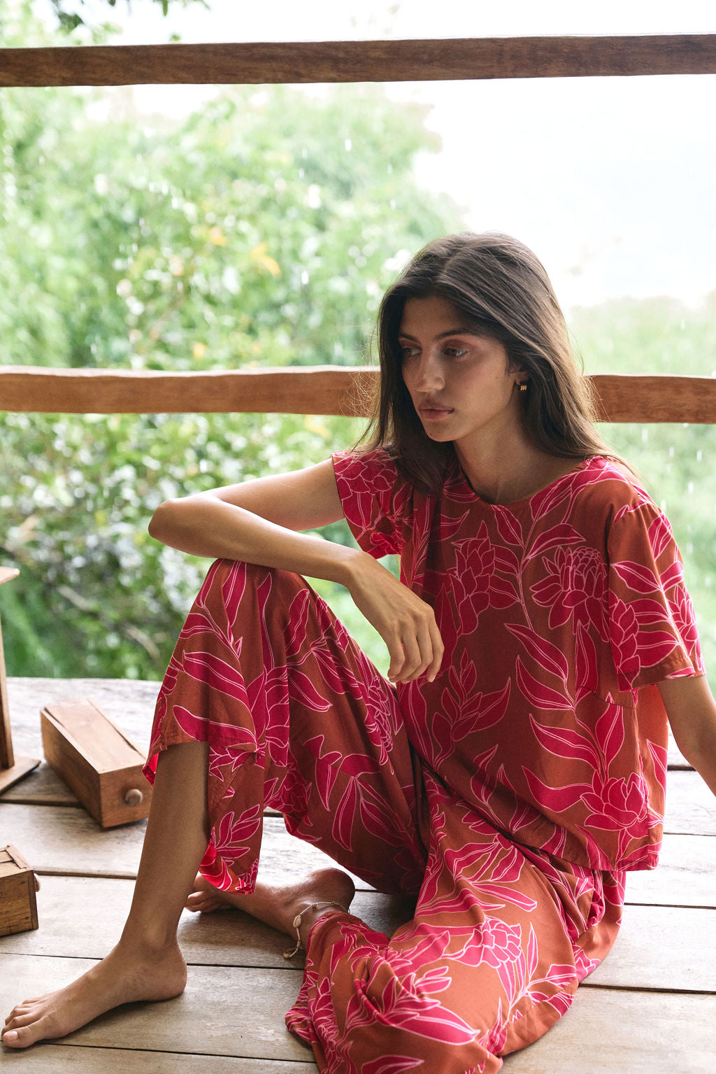 A woman with dark hair wears a YIREH t-shirt top with shell button back. It is the Noe Top in the Garden 'Awapuhi print.