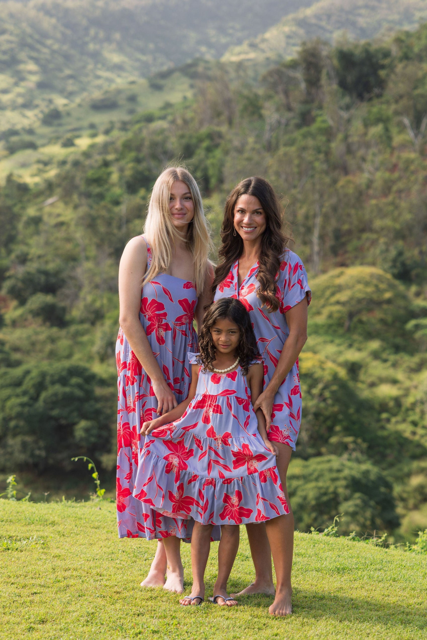little girl with brown hair wearing a mid length dress with periwinkle background and bright red hibiscus print.
