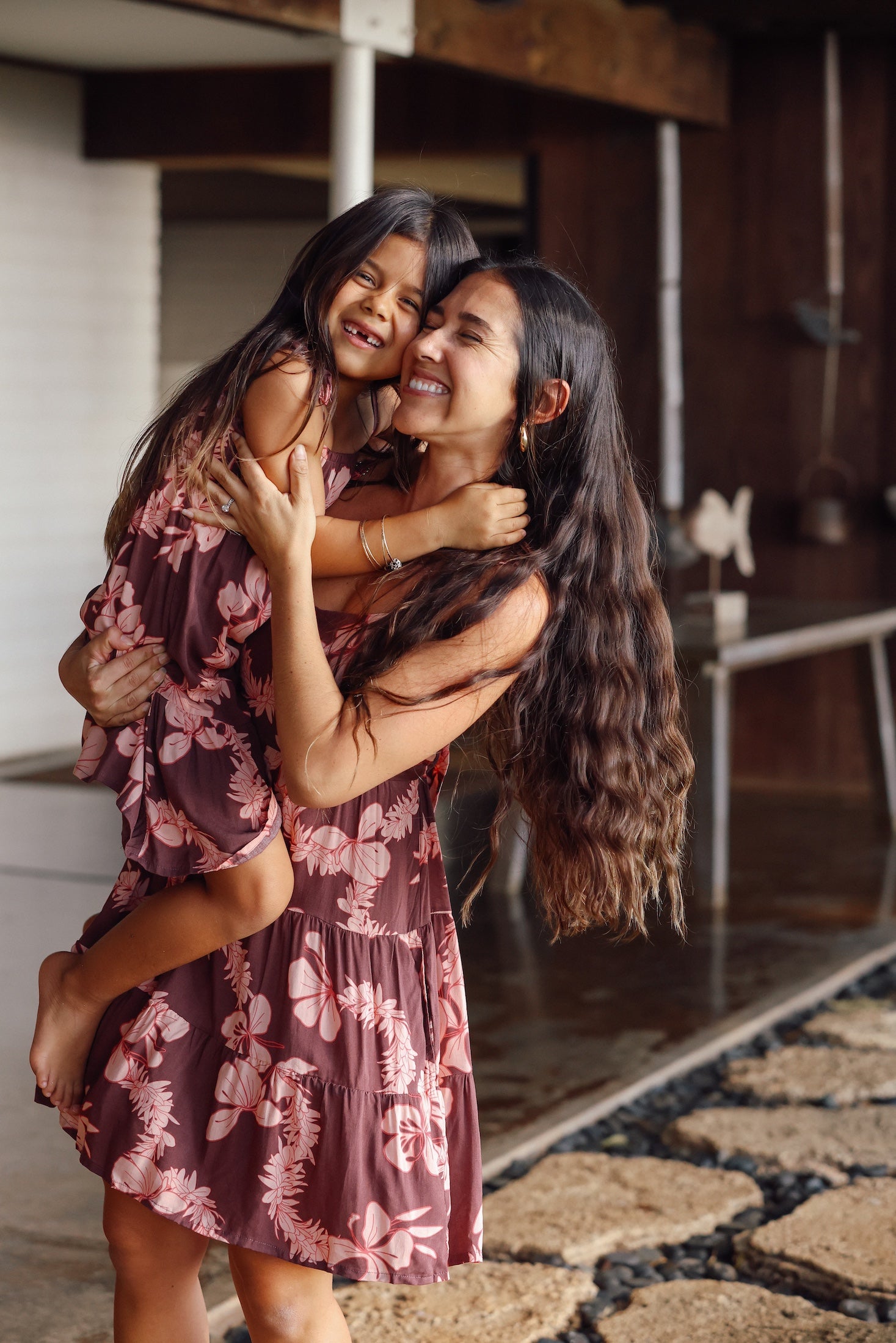 little girl wearing a long brown dress with tie sleeve with light pink lei awapuhi print hugging her mother in matching dress