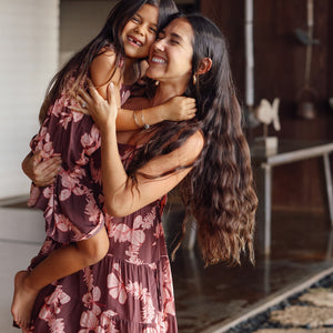 little girl wearing a long brown dress with tie sleeve with light pink lei awapuhi print hugging her mother in matching dress