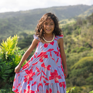 little girl with brown hair wearing a mid length dress with periwinkle background and bright red hibiscus print.