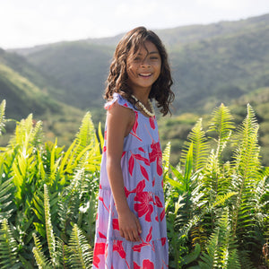 little girl with brown hair wearing a mid length dress with periwinkle background and bright red hibiscus print.