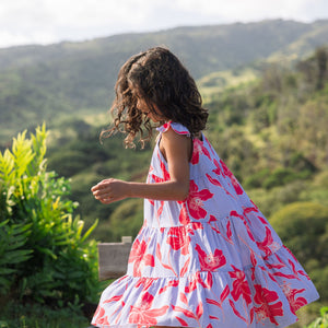 little girl with brown hair wearing a mid length dress with periwinkle background and bright red hibiscus print.