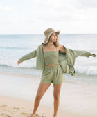 Woman wearing loose button up with smocked tube top and short in medium green linen.