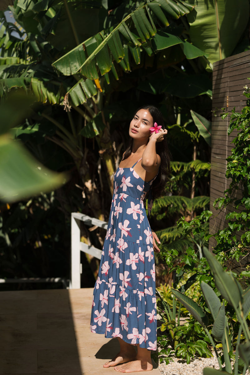 woman with black hair wearing a maxi tiered dress in a navy color with pink hibiscus blossoms 