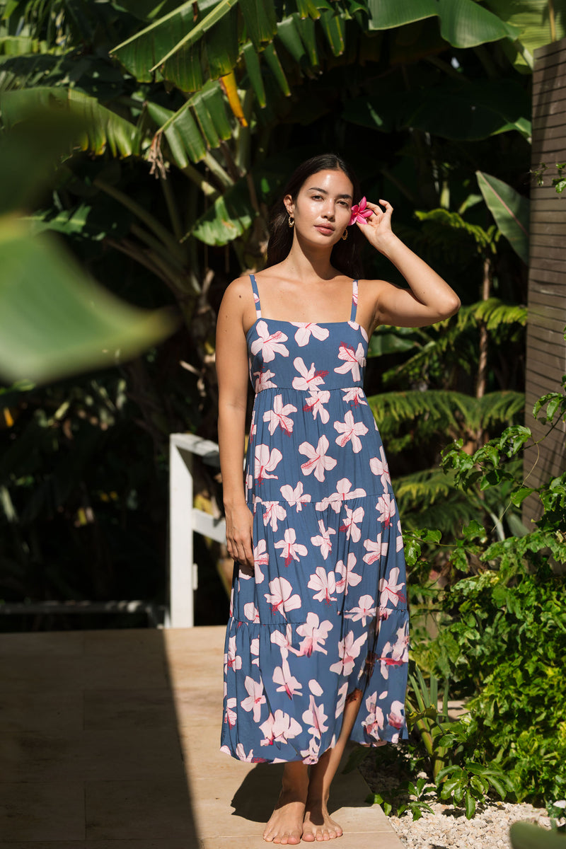woman with black hair wearing a maxi tiered dress in a navy color with pink hibiscus blossoms 