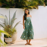woman with brown hair wearing lush green maxi spaghetti strap dress with a tie back detail and puakenikeni print