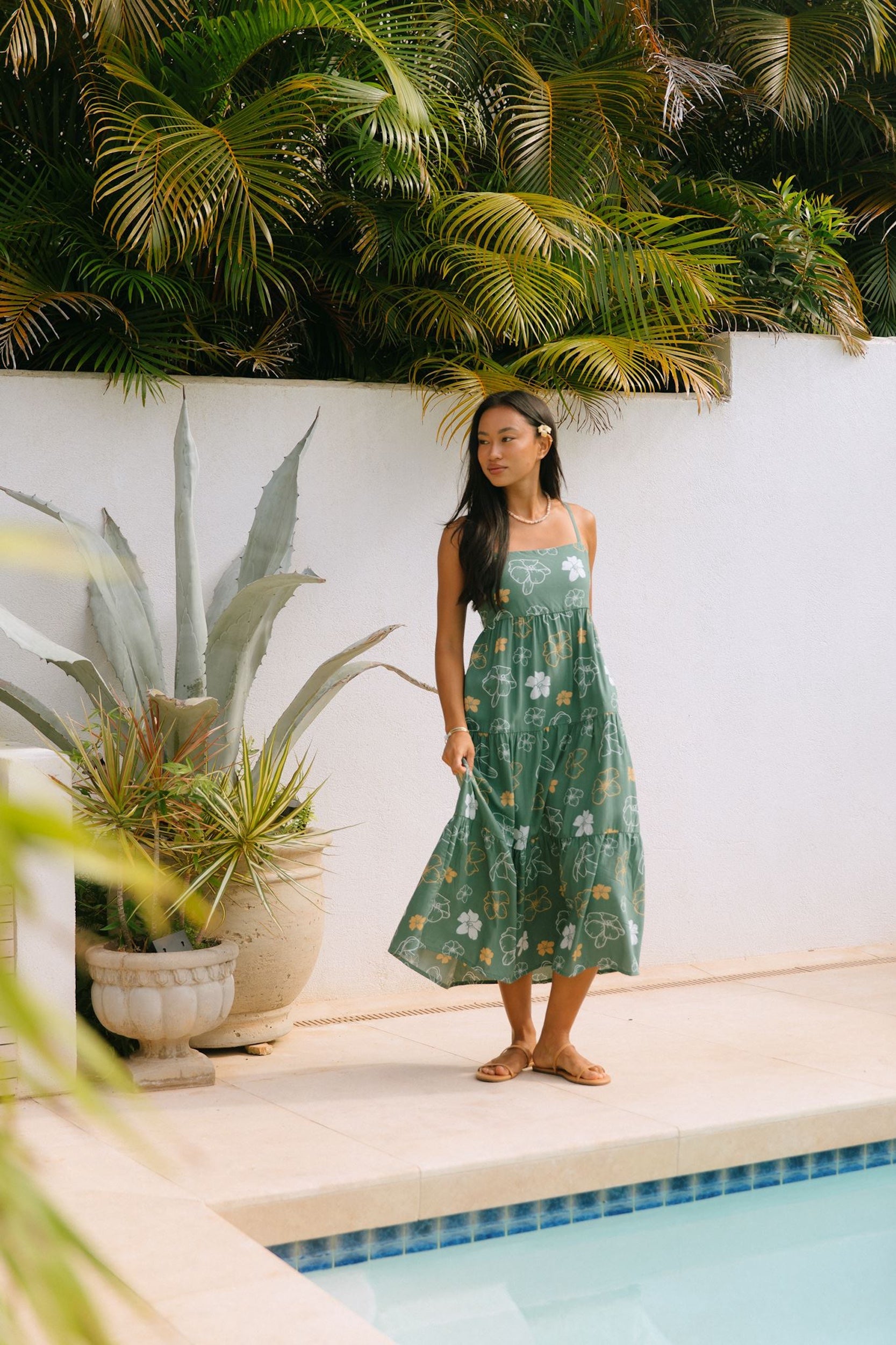 woman with brown hair wearing lush green maxi spaghetti strap dress with a tie back detail and puakenikeni print