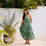 woman with brown hair wearing lush green maxi spaghetti strap dress with a tie back detail and puakenikeni print