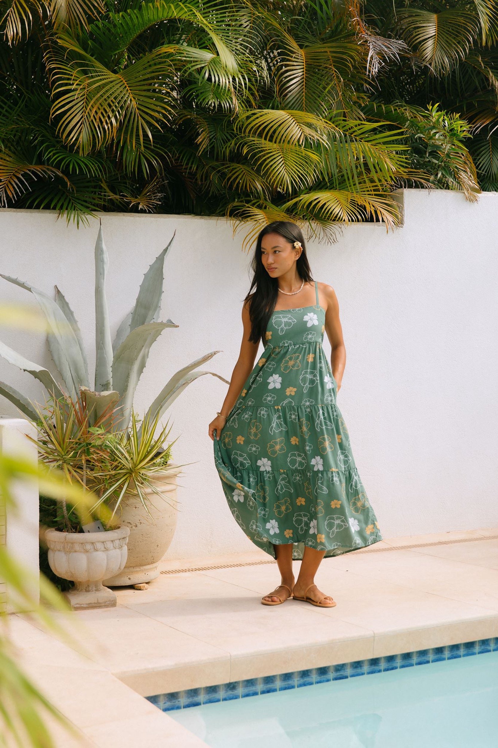 woman with brown hair wearing lush green maxi spaghetti strap dress with a tie back detail and puakenikeni print