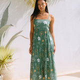 woman with brown hair wearing lush green maxi spaghetti strap dress with a tie back detail and puakenikeni print