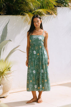 woman with brown hair wearing lush green maxi spaghetti strap dress with a tie back detail and puakenikeni print