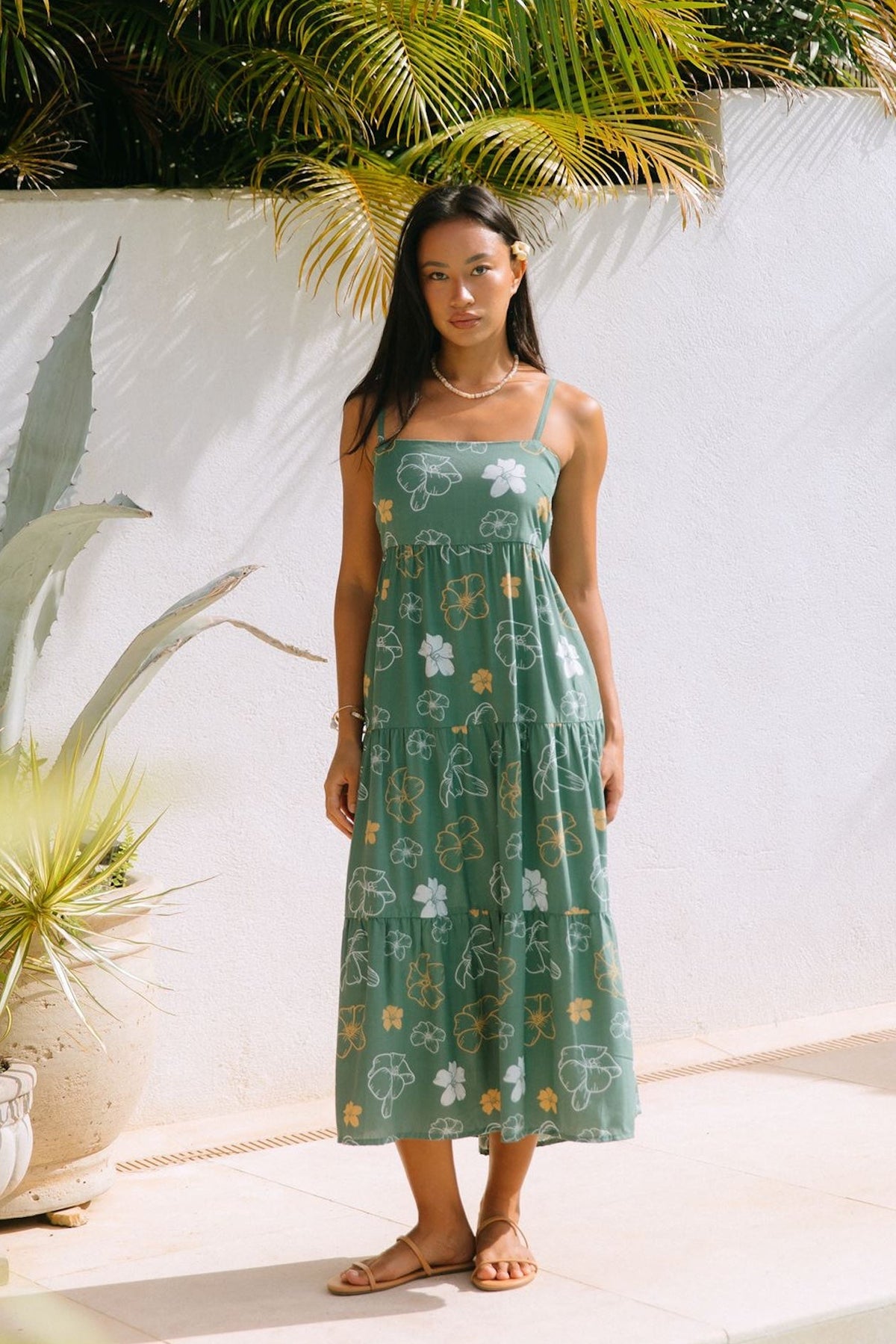 woman with brown hair wearing lush green maxi spaghetti strap dress with a tie back detail and puakenikeni print