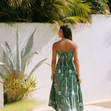 woman with brown hair wearing lush green maxi spaghetti strap dress with a tie back detail and puakenikeni print