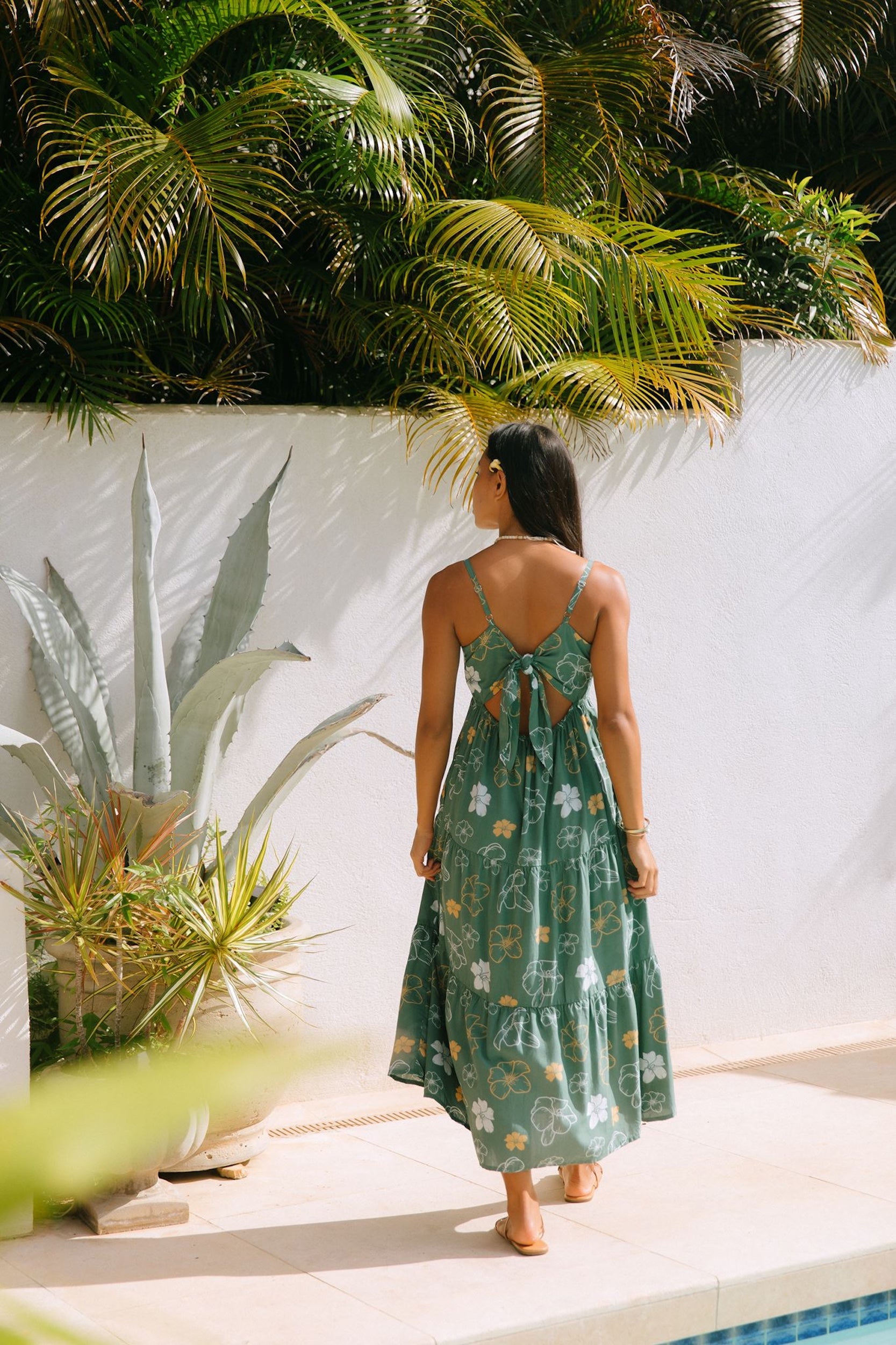 woman with brown hair wearing lush green maxi spaghetti strap dress with a tie back detail and puakenikeni print