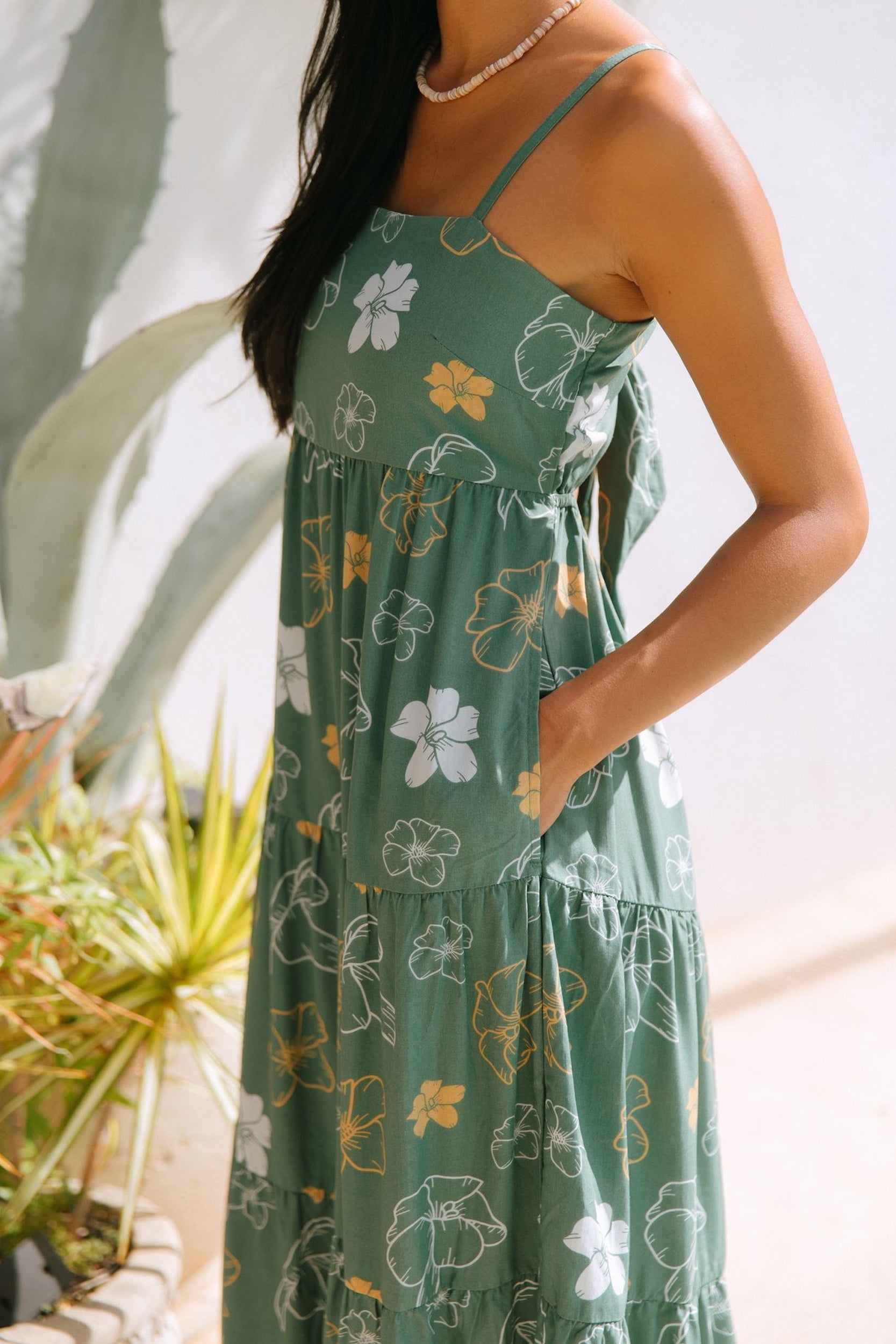 woman with brown hair wearing lush green maxi spaghetti strap dress with a tie back detail and puakenikeni print