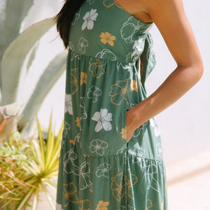 woman with brown hair wearing lush green maxi spaghetti strap dress with a tie back detail and puakenikeni print