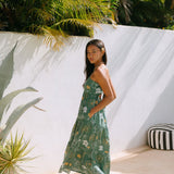 woman with brown hair wearing lush green maxi spaghetti strap dress with a tie back detail and puakenikeni print