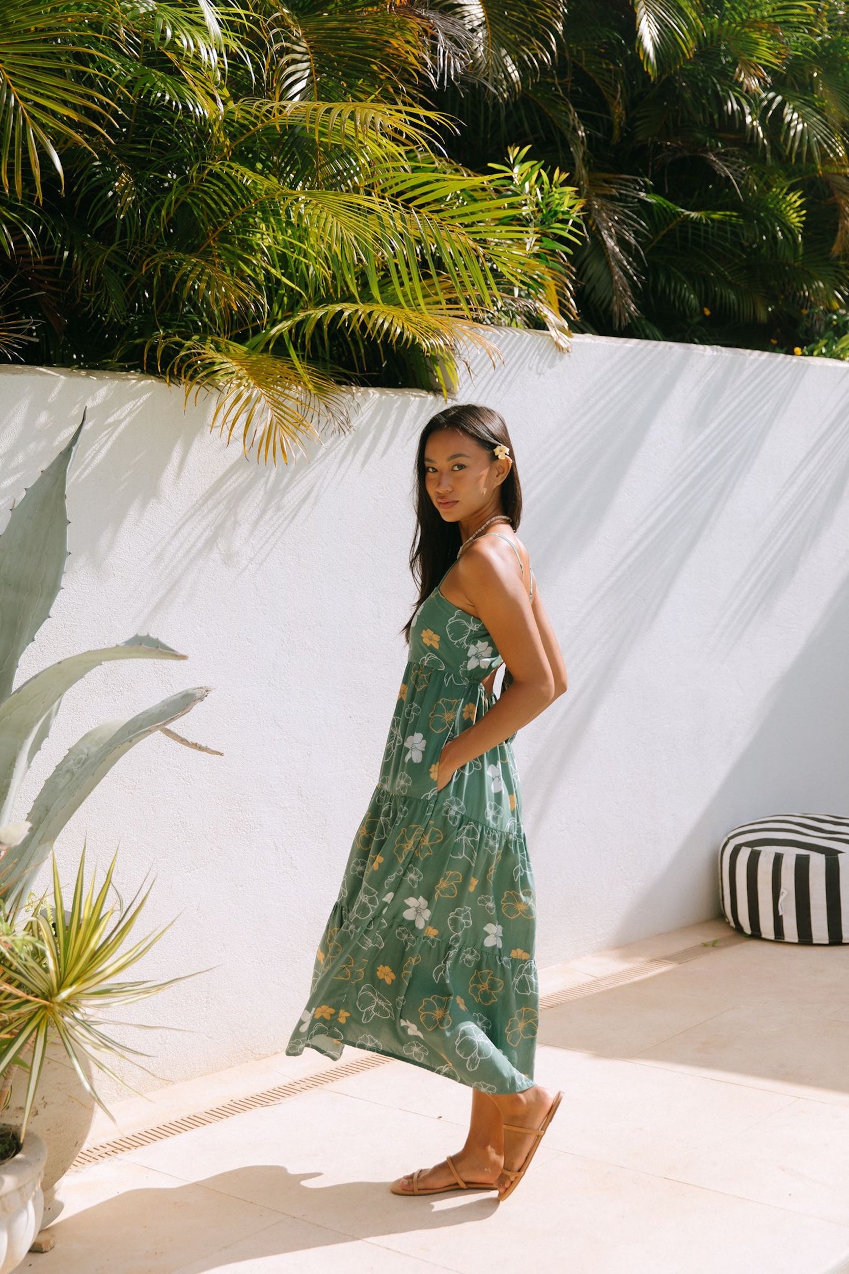 woman with brown hair wearing lush green maxi spaghetti strap dress with a tie back detail and puakenikeni print