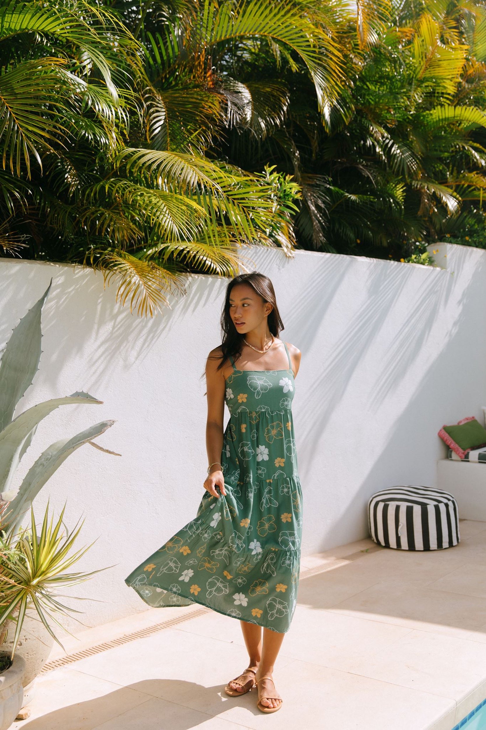 woman with brown hair wearing lush green maxi spaghetti strap dress with a tie back detail and puakenikeni print