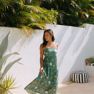 woman with brown hair wearing lush green maxi spaghetti strap dress with a tie back detail and puakenikeni print