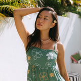 woman with brown hair wearing lush green maxi spaghetti strap dress with a tie back detail and puakenikeni print