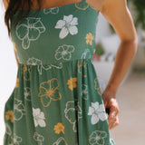 woman with brown hair wearing lush green maxi spaghetti strap dress with a tie back detail and puakenikeni print