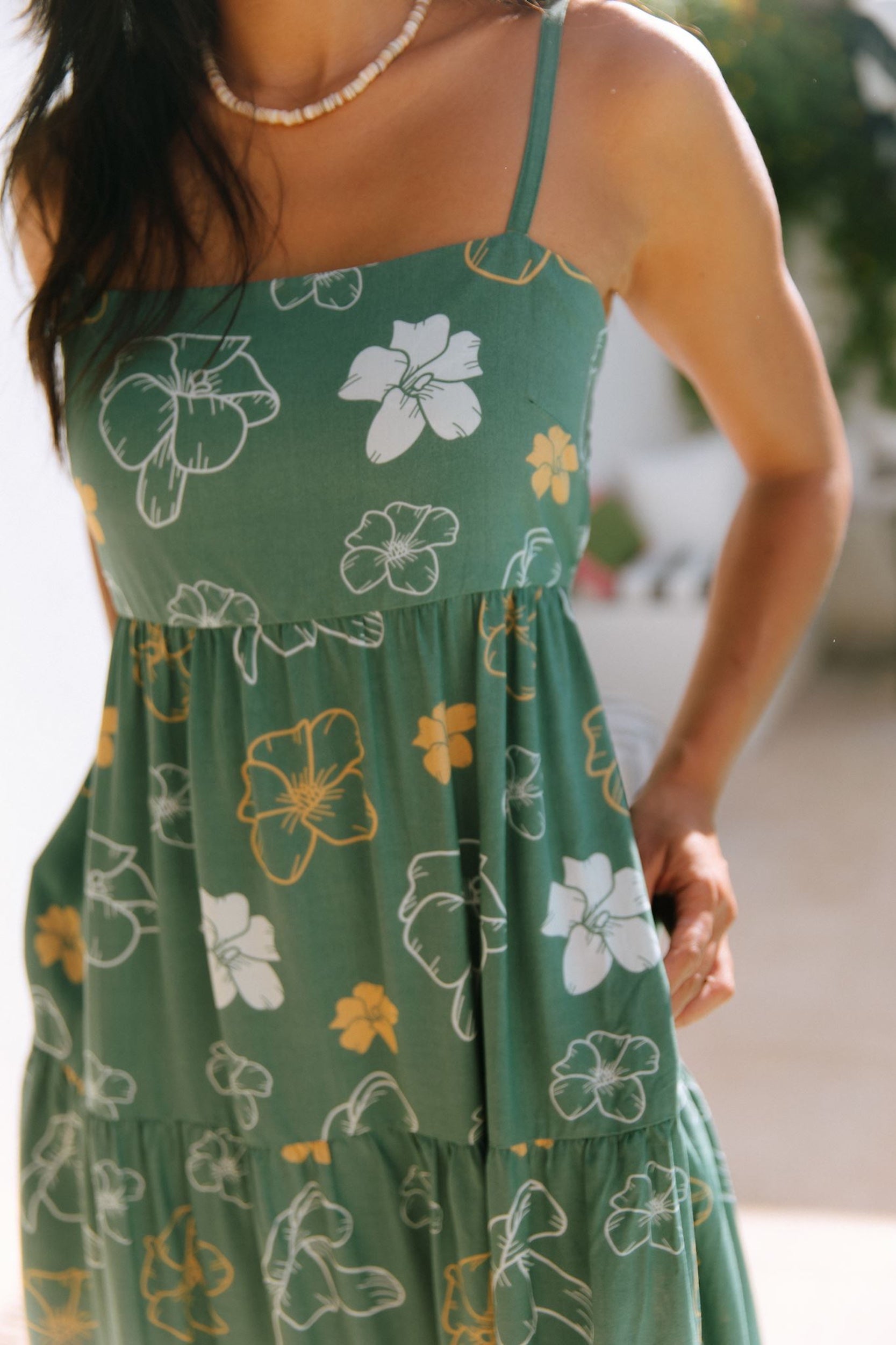 woman with brown hair wearing lush green maxi spaghetti strap dress with a tie back detail and puakenikeni print