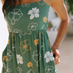 woman with brown hair wearing lush green maxi spaghetti strap dress with a tie back detail and puakenikeni print