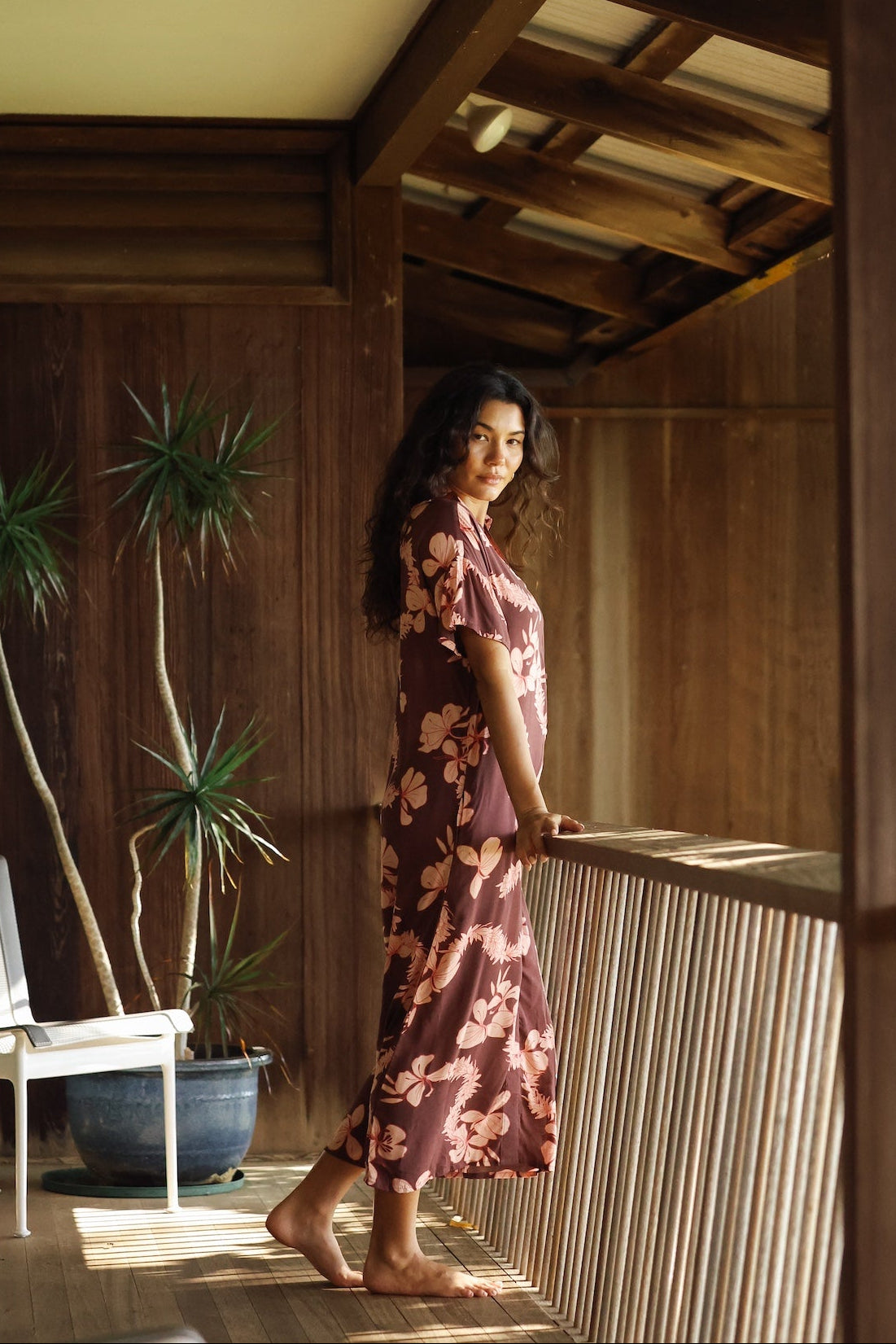 woman with brown hair wearing a brown colored maxi button up dress with light pink lei awapuhi print