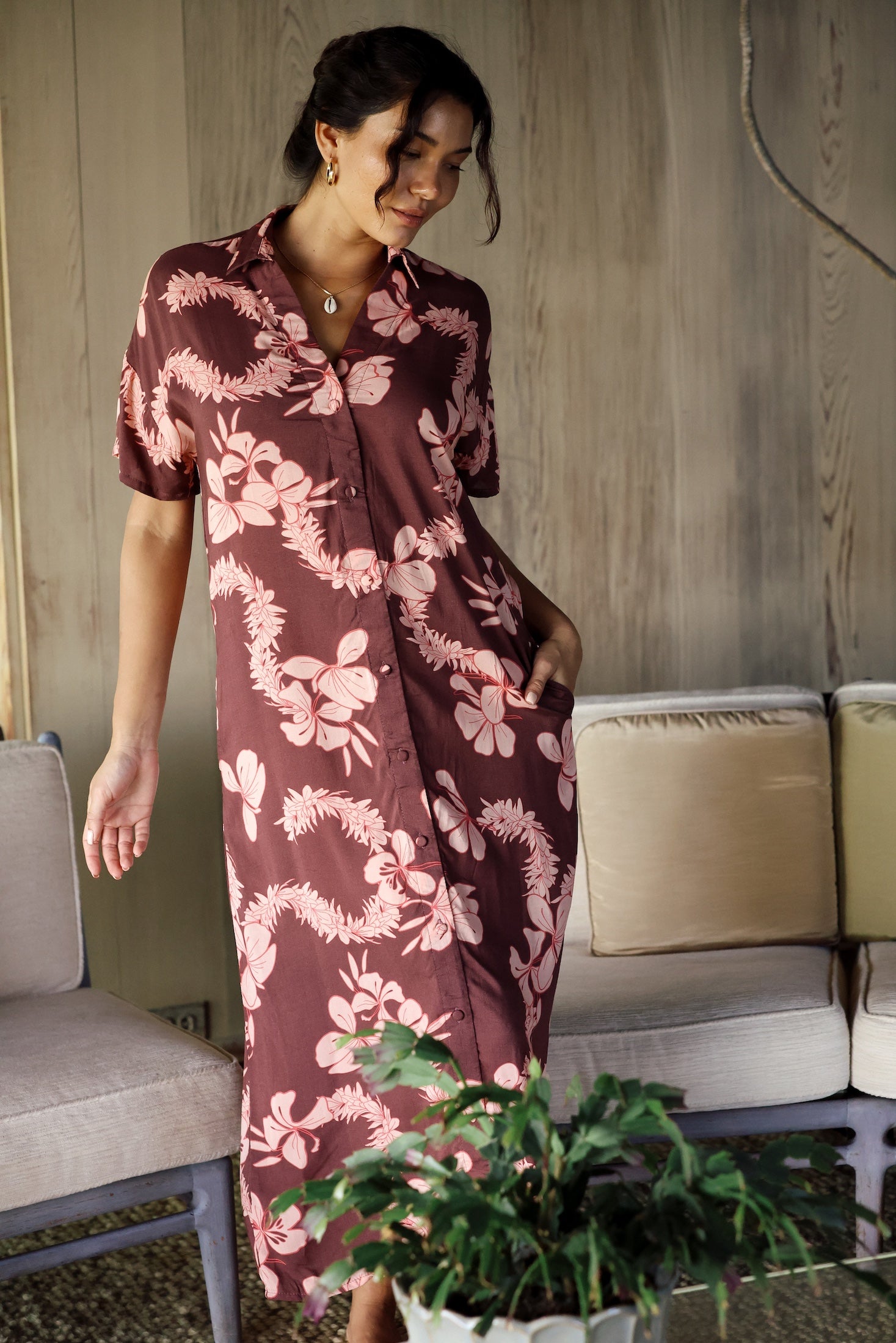 woman with brown hair wearing a brown colored maxi button up dress with light pink lei awapuhi print