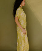 Woman in long short sleeve shirt dress with beige floral print on light green ground.