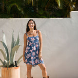 woman with black hair wearing a mini tiered dress with tie back in a navy color with pink hibiscus blossoms 