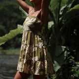 woman with brown hair wearing a mini dress with a tie back in a green color with naupaka pattern throughout. 