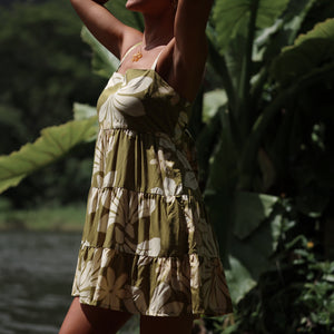 woman with brown hair wearing a mini dress with a tie back in a green color with naupaka pattern throughout. 