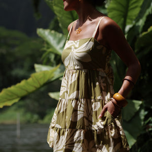 woman with brown hair wearing a mini dress with a tie back in a green color with naupaka pattern throughout. 