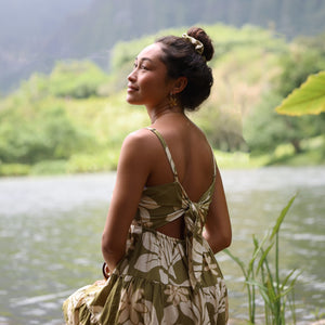 woman with brown hair wearing a mini dress with a tie back in a green color with naupaka pattern throughout. 