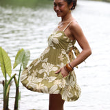 woman with brown hair wearing a mini dress with a tie back in a green color with naupaka pattern throughout. 