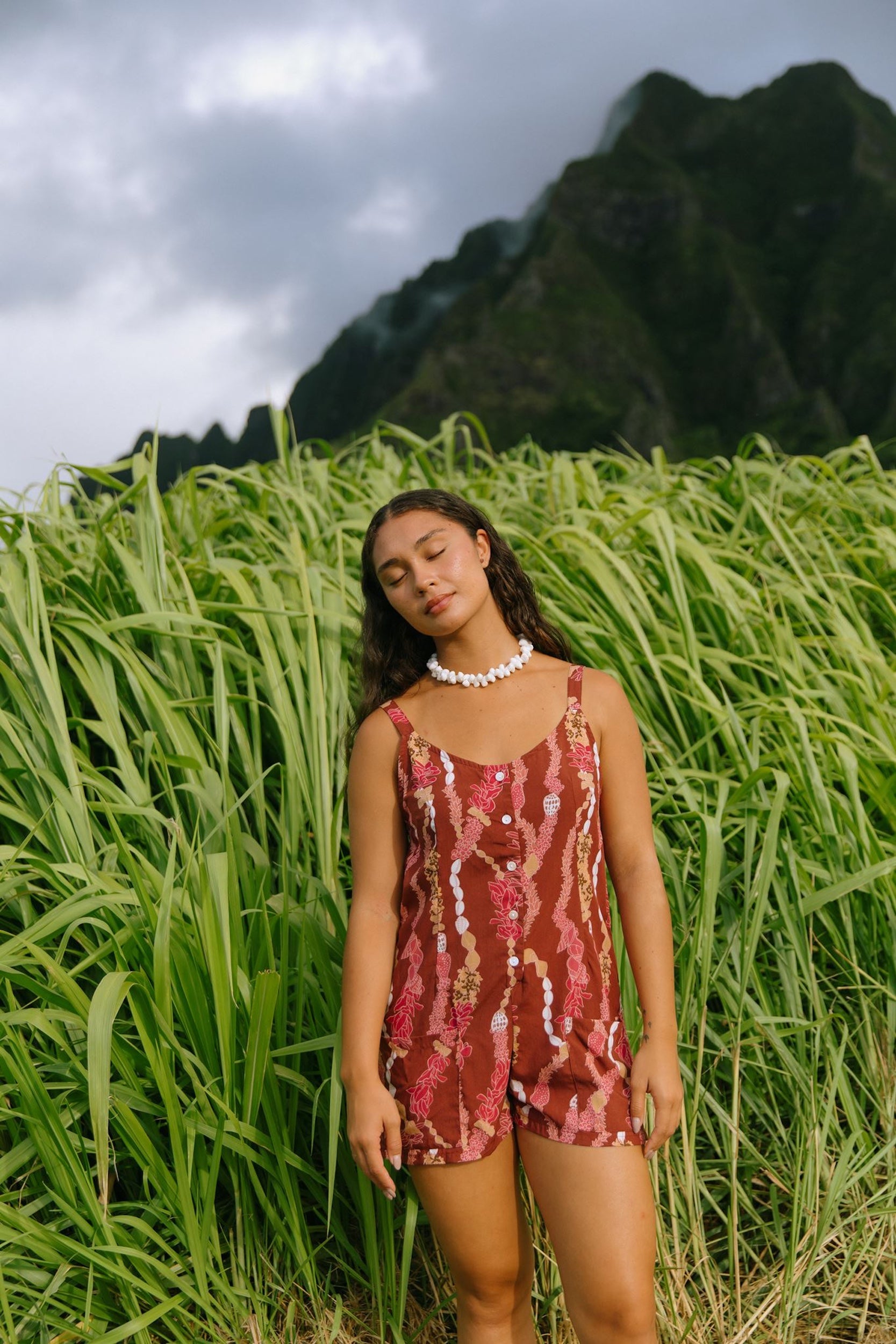 woman with brown hair wearing a red-brown colored romper with buttons, a tie back, front pockets, and lei print
