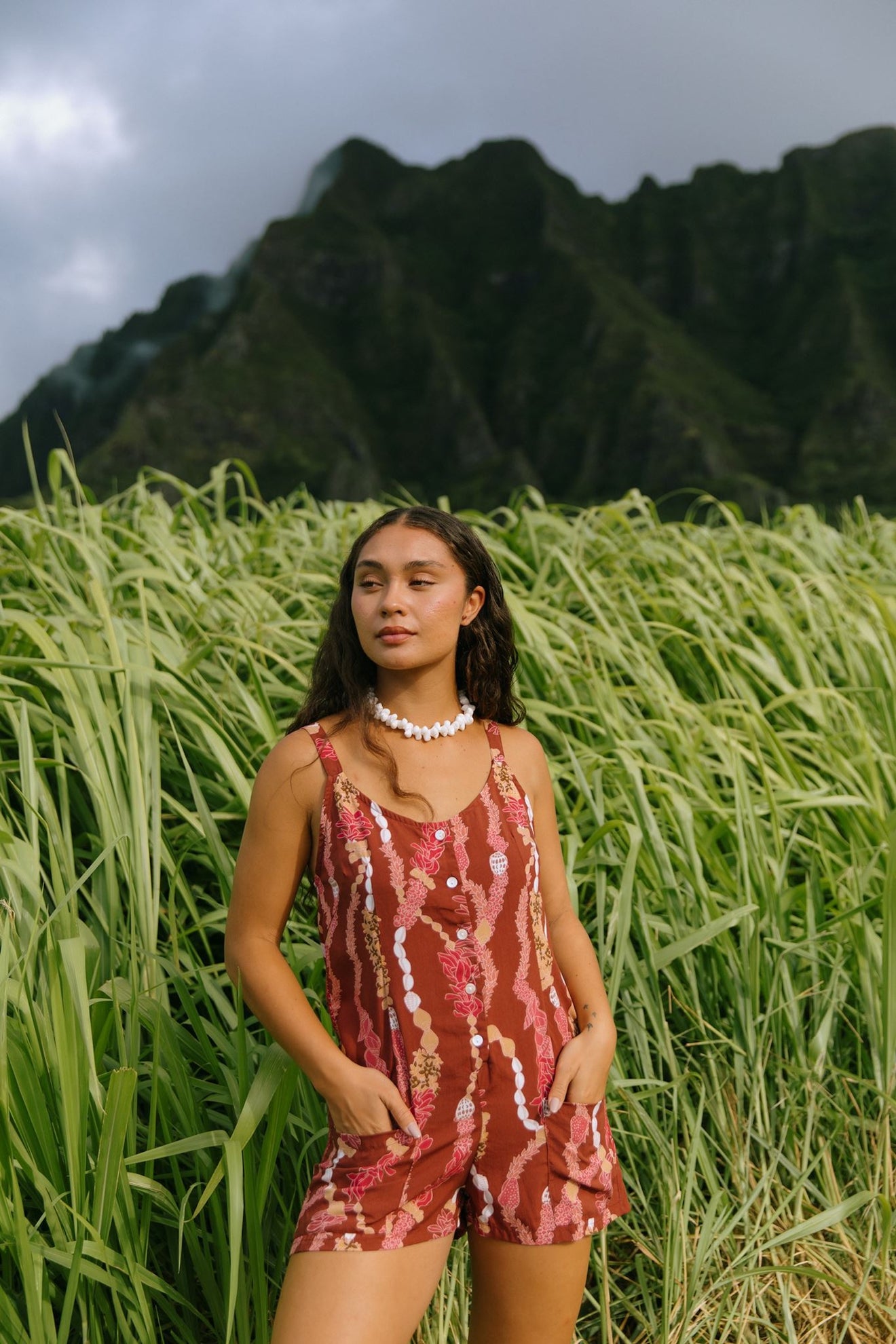 woman with brown hair wearing a red-brown colored romper with buttons, a tie back, front pockets, and lei print