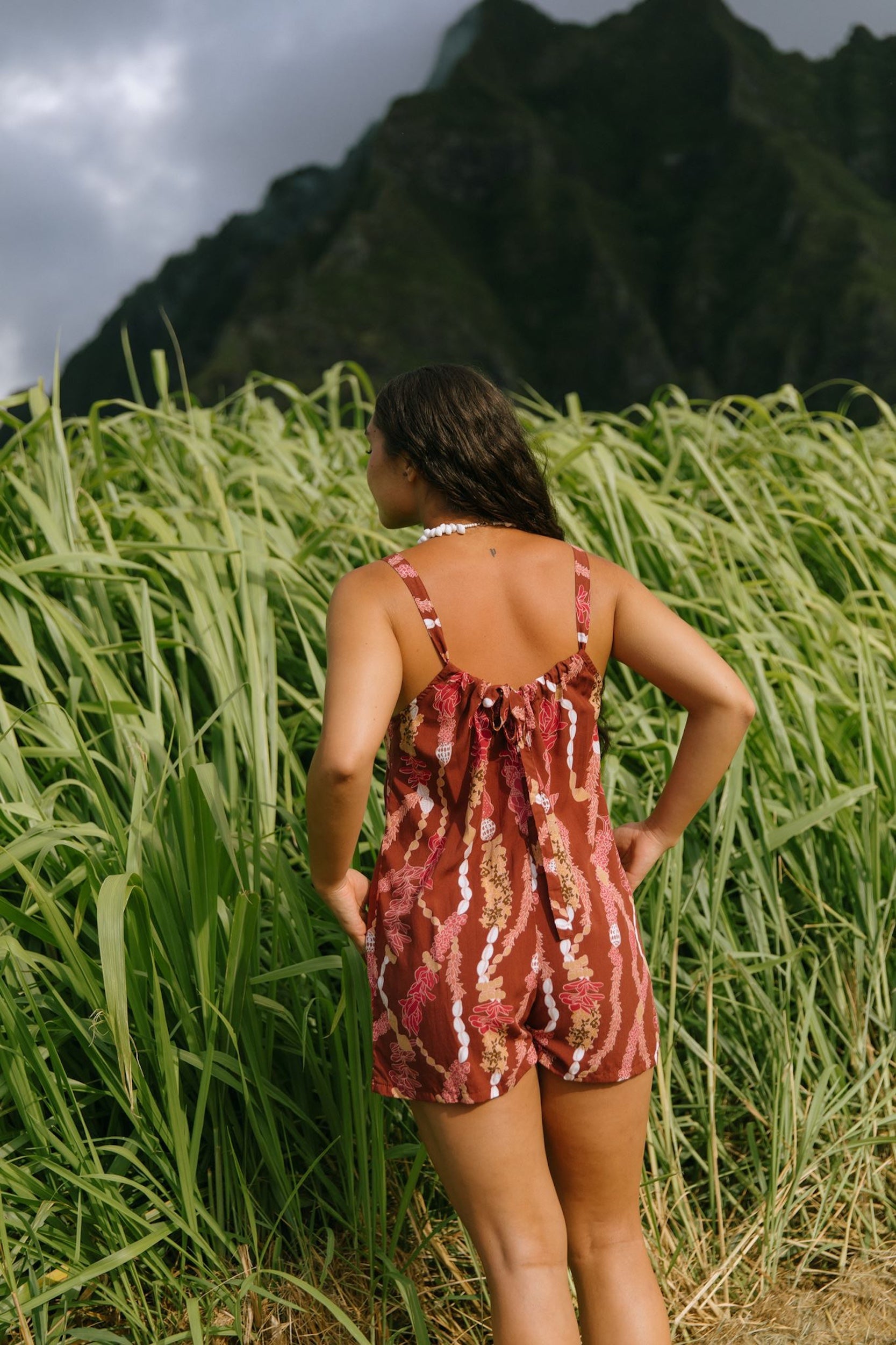 woman with brown hair wearing a red-brown colored romper with buttons, a tie back, front pockets, and lei print