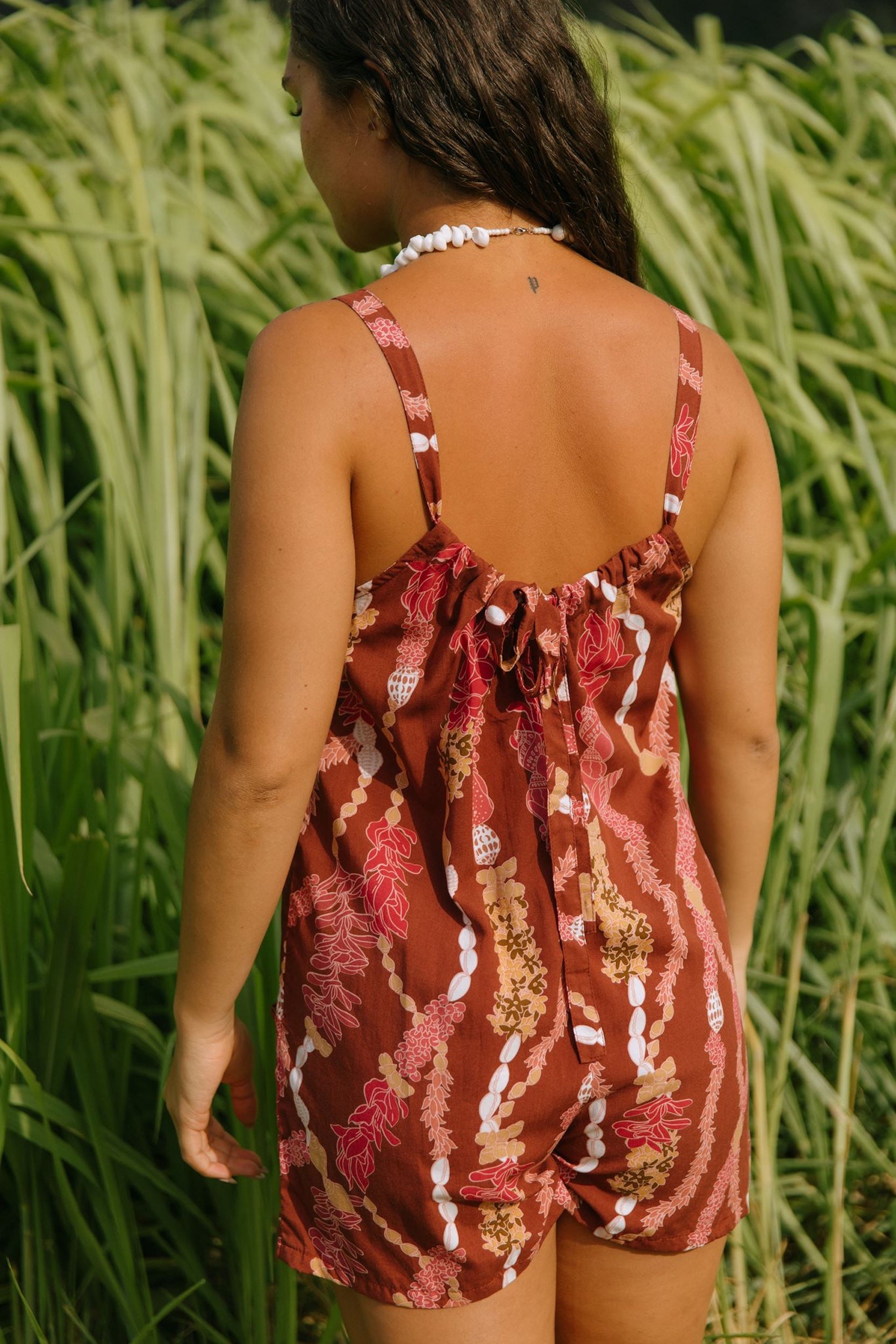 woman with brown hair wearing a red-brown colored romper with buttons, a tie back, front pockets, and lei print